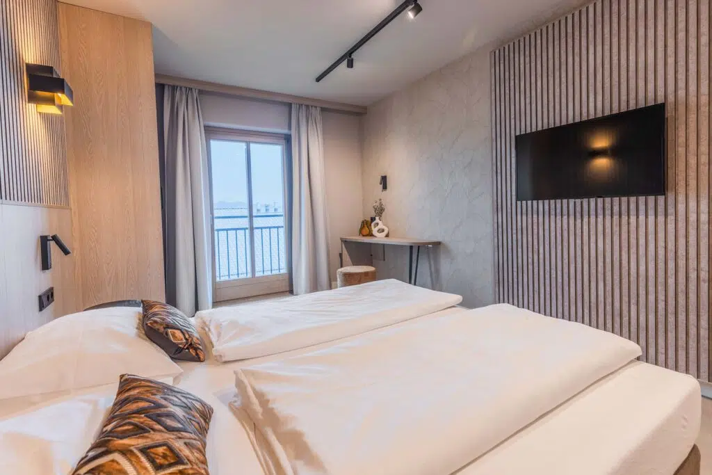 Zweibettzimmer mit gemusterten Kissen, Lamellenwand und Blick auf die Dächer, Aldiana Club Ampflwang