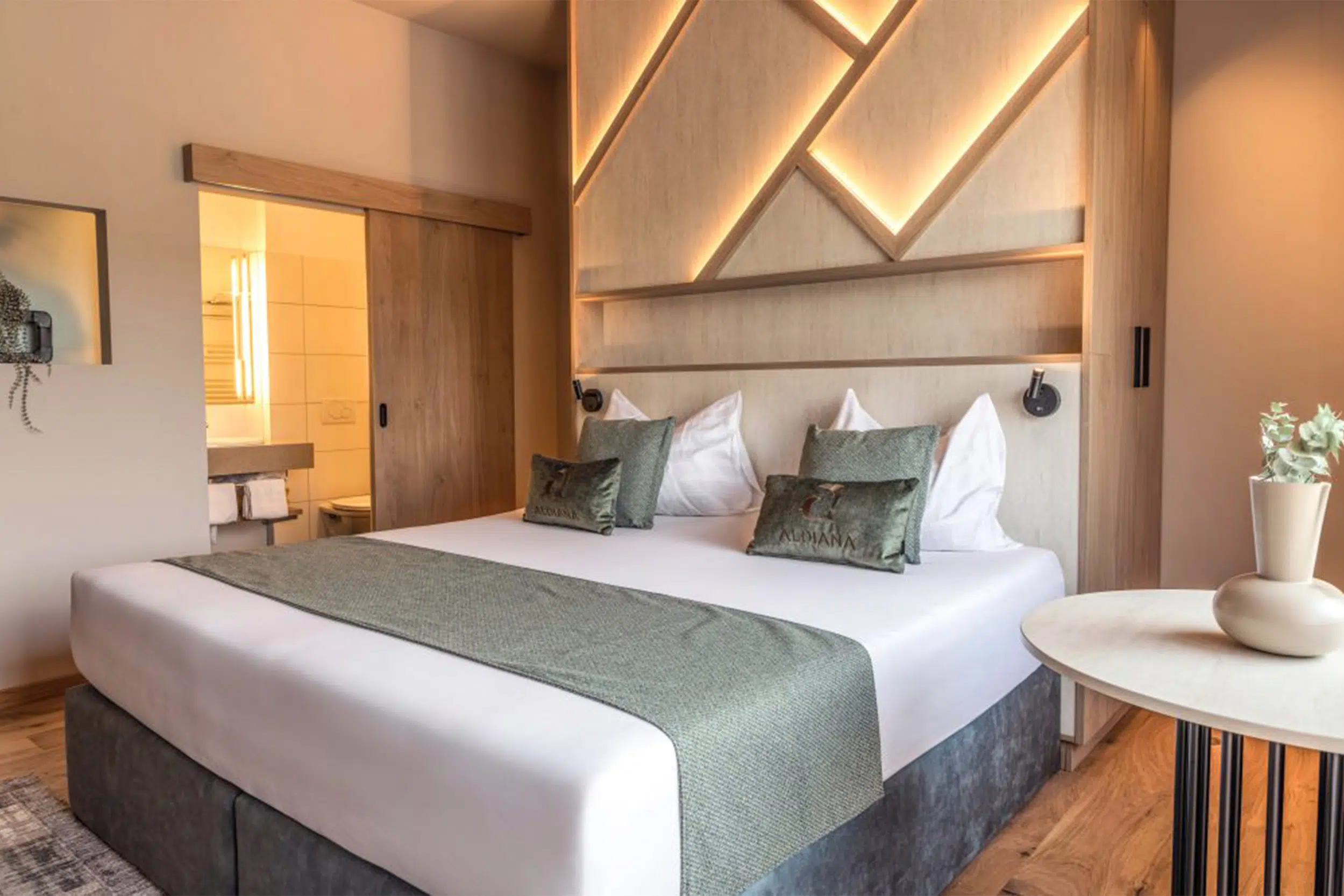Doppelbett vor beleuchteter Bergsilhouette aus Holz, grüne Kissen und Schiebetür zum Bad, Aldiana Club Salzkammergut