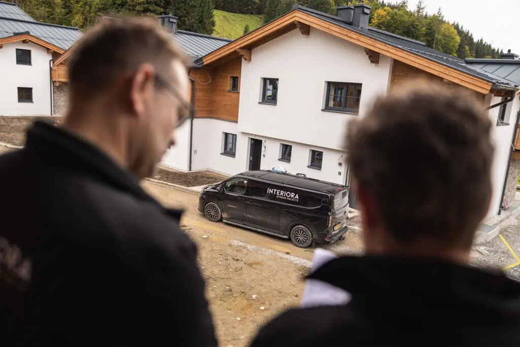 Blick über die Schulter zweier Mitarbeiter auf den Firmenbus vor einem Chalet, Tauernlodge